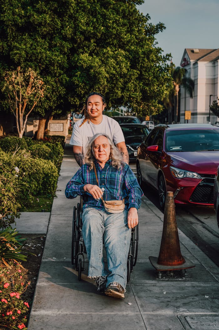 A caring scene of a young man pushing a senior in a wheelchair along a sidewalk.