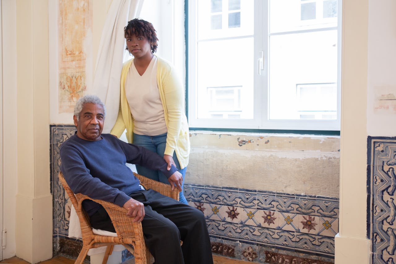 Elderly man seated with caregiver standing beside him at home in Portugal.